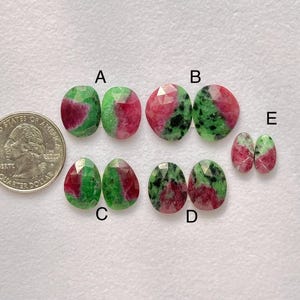 Könnte beinhalten: Auswahl an facettierten Rubin-Zoisit-Edelsteinen in verschiedenen Formen, darunter oval und tropfenförmig. Die Steine zeigen eine Mischung aus grünen und roten Farbtönen, wobei einige schwarze Einschlüsse aufweisen. Eine amerikanische Vierteldollarmünze dient als Größenreferenz.