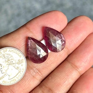Può includere: Due pietre preziose a forma di pera, sfaccettate, di un colore rosso rosato intenso con una finitura lucida. Una moneta da venticinque centesimi di dollaro statunitense è posizionata accanto alle pietre per fornire un riferimento visivo.