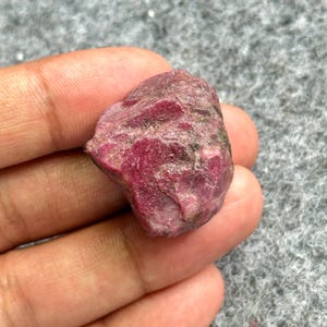 May include: A rough, uncut ruby gemstone with a deep red hue. The natural stone has an irregular shape and a textured surface, showcasing its raw, unpolished state. The ruby is held in a hand against a gray background.