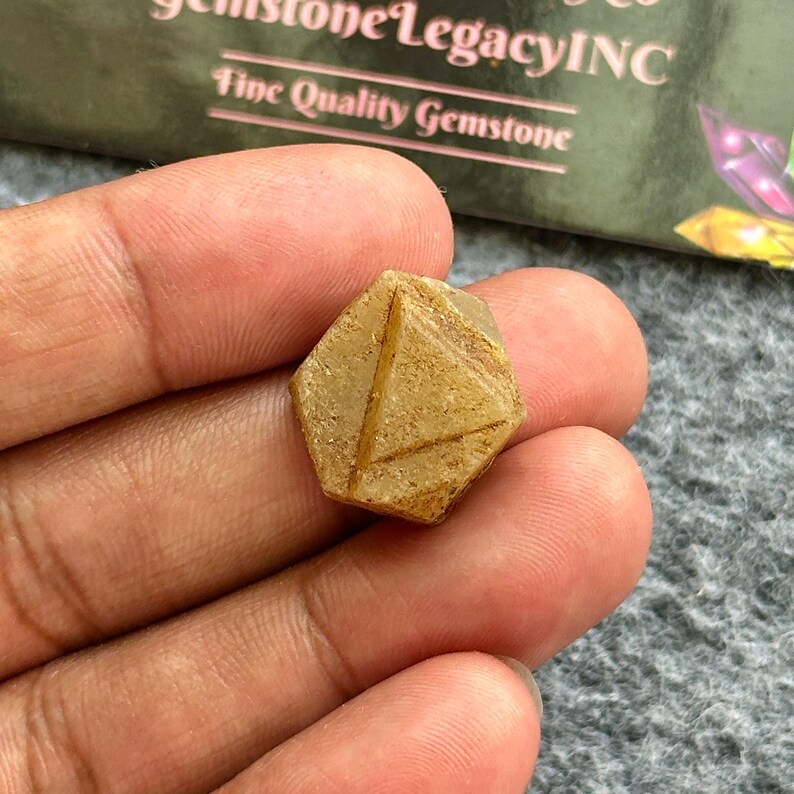 Op de afbeelding: Een gefacetteerde, lichtbruine edelsteen in een hand. De edelsteen heeft een geometrische vorm met zichtbare lijnen en facetten. De achtergrond bevat de tekst "Gemstone Legacy INC" en "Fine Quality Gemstone".