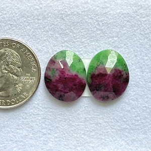 May include: Two oval-shaped, faceted ruby zoisite gemstones featuring a vibrant mix of deep magenta and bright green colors. The stones are positioned next to a United States quarter coin, which has a diameter of approximately 0.955 inches.