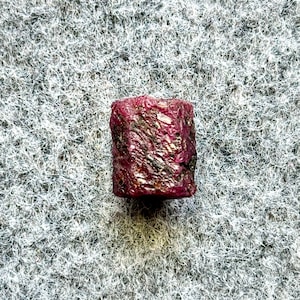 May include: A close-up of a dark red ruby gemstone. The ruby is faceted and has a slightly rough texture. It is set against a white, fuzzy background.