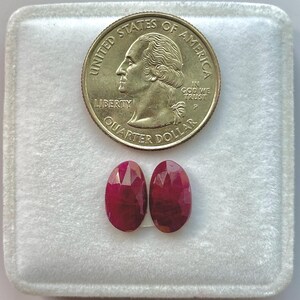 Puede incluir: Dos rubíes rojos facetados de forma ovalada colocados debajo de una moneda de veinticinco centavos de dólar estadounidense. Los rubíes muestran un tono carmesí profundo y una superficie pulida, ofreciendo una referencia de escala clara frente a la moneda de 2,43 cm.