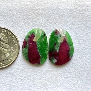 Puede incluir: Dos gemas facetadas de forma ovalada con una mezcla de colores verde y rojo. Las gemas están junto a una moneda de veinticinco centavos para comparar el tamaño. Las gemas tienen una superficie pulida y reflectante.