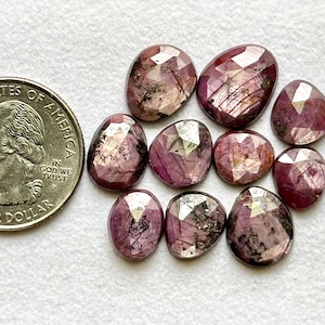 Peut inclure: Plusieurs pierres précieuses rubis ovales et facettées de différentes tailles, présentant une teinte rouge profond avec des inclusions internes. Une pièce de monnaie américaine est incluse pour l'échelle, sur fond blanc.