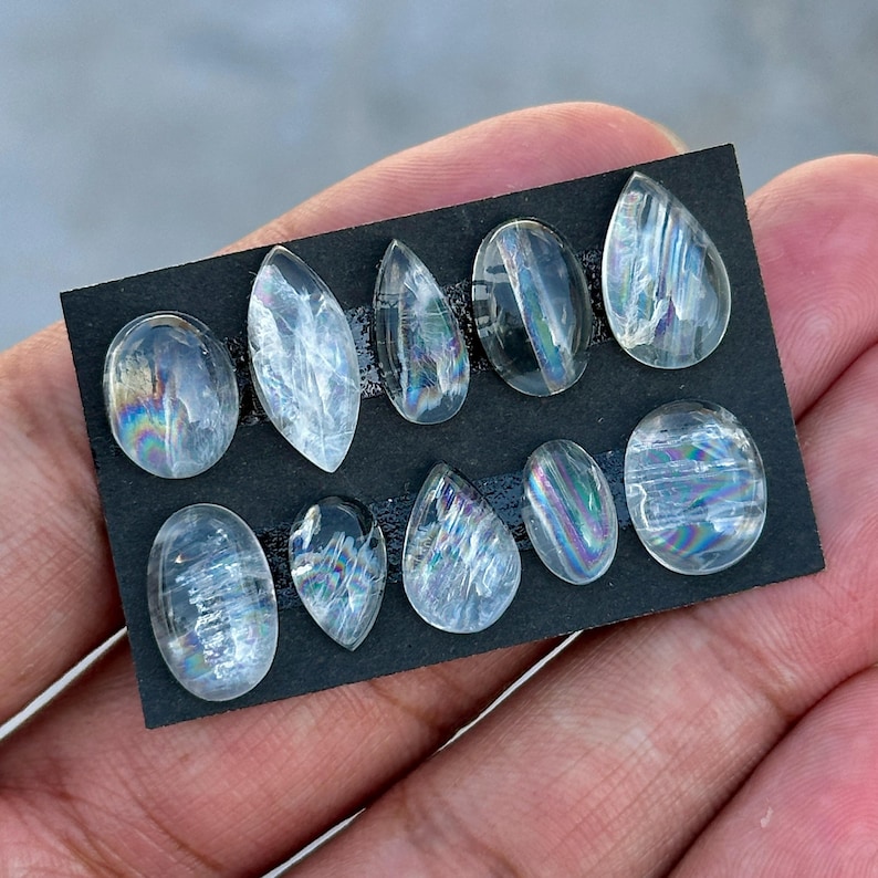 May include: A collection of ten clear, iridescent gemstones in various shapes, including oval, teardrop, and pointed designs. The gemstones are arranged on a black rectangular card, showcasing their internal rainbow-like patterns and clarity.