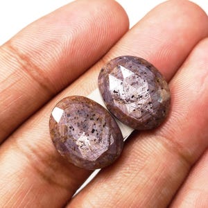 Pode incluir: Duas pedras preciosas ovais facetadas de cor roxa e marrom. As pedras têm uma qualidade brilhante e iridescente.