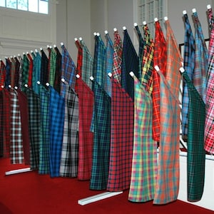 May include: A display of various tartan fabrics hanging on white poles. The fabrics are in a variety of colours, including red, green, blue, and yellow. The fabrics are arranged in a row, with the poles extending out from the wall.