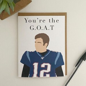 May include: A white card with brown trim features a cartoon illustration of a football player wearing a blue jersey with the number 12. The text "You're the G.O.A.T." is printed above the illustration.