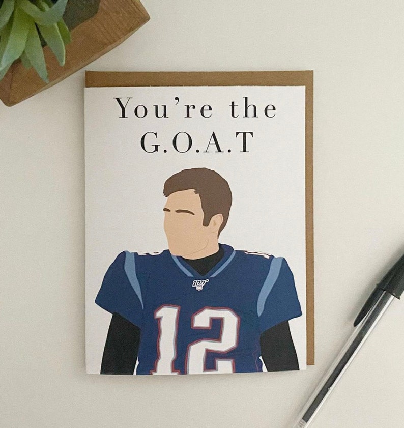 May include: A white card with brown trim features a cartoon illustration of a football player wearing a blue jersey with the number 12. The text "You're the G.O.A.T." is printed above the illustration.