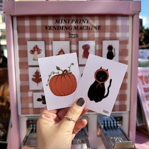 May include: Two rectangular white cards featuring illustrations of a pumpkin and a black cat wearing an orange bonnet. The cards are displayed in front of a pink mini print vending machine with a light pink and white gingham background pattern.