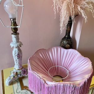 May include: A vintage lamp with a white porcelain base decorated with flowers and a pink fringed lampshade. The lamp has a white metal frame and a lightbulb. The lampshade is approximately 30 cm in diameter.
