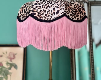 May include: A lampshade featuring a leopard print design, black trim, and pink fringe. The lampshade is mounted on a gold-coloured lamp stand. The background is teal.