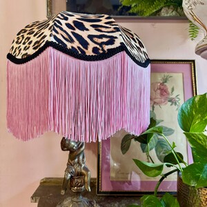 May include: A decorative table lamp featuring a leopard print lampshade and pink fringe. The lamp has a bronze-coloured base with a cherub figure. The lamp is on a dark surface.