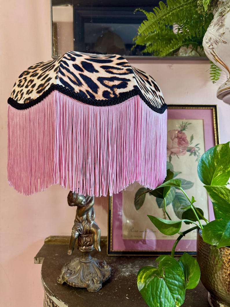 May include: A decorative table lamp featuring a leopard print lampshade and pink fringe. The lamp has a bronze-coloured base with a cherub figure. The lamp is on a dark surface.