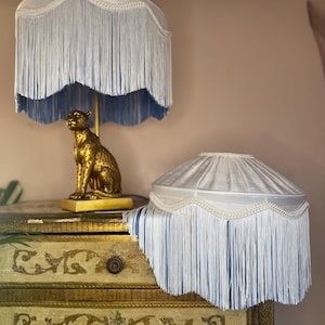 Può includere: Due paralumi con frange blu chiaro con una base a statua di leopardo dorato poggiano su un comò vintage. I paralumi hanno un bordo smerlato e finiture color crema. Il comò presenta dettagli dorati ornati.