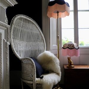 May include: A white wicker peacock chair with a sheepskin throw and a navy blue cushion. A gold floor lamp with a fringed lampshade and a leopard print table lamp sit near a window. The lampshades have a black trim.