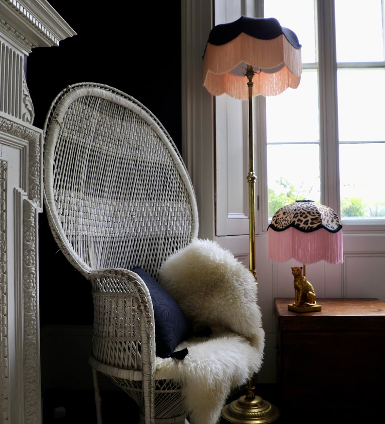 May include: A white wicker peacock chair with a sheepskin throw and a navy blue cushion. A gold floor lamp with a fringed lampshade and a leopard print table lamp sit near a window. The lampshades have a black trim.