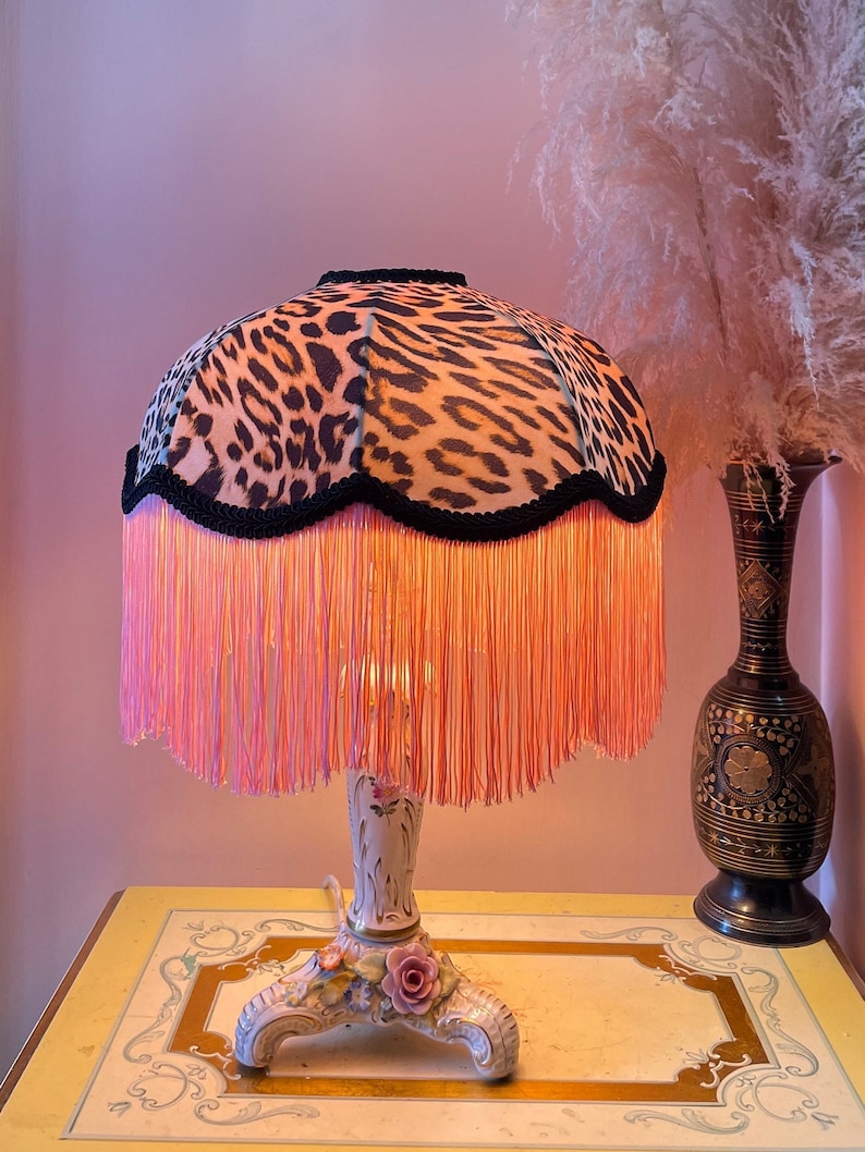 May include: A table lamp featuring a leopard print lampshade with pink fringe. The base is white with floral details. The lamp sits on a yellow table with decorative trim. A vase with tall, feathery plumes is to the right.