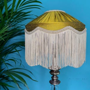 Può includere: Lampada da tavolo in stile vintage con paralume plissettato verde oliva. Il paralume è rifinito con frange bianche e finiture decorative. La base della lampada è una combinazione di vetro trasparente e metallo. Sfondo turchese.