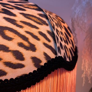 May include: A lampshade with a leopard print pattern in brown and tan. The shade is trimmed with black velvet and has long, peach-coloured fringe.