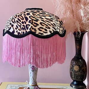 May include: A decorative table lamp with a leopard print lampshade, trimmed with black rope and pink fringe. The base is white porcelain with floral accents. A black vase with gold detailing is to the side.
