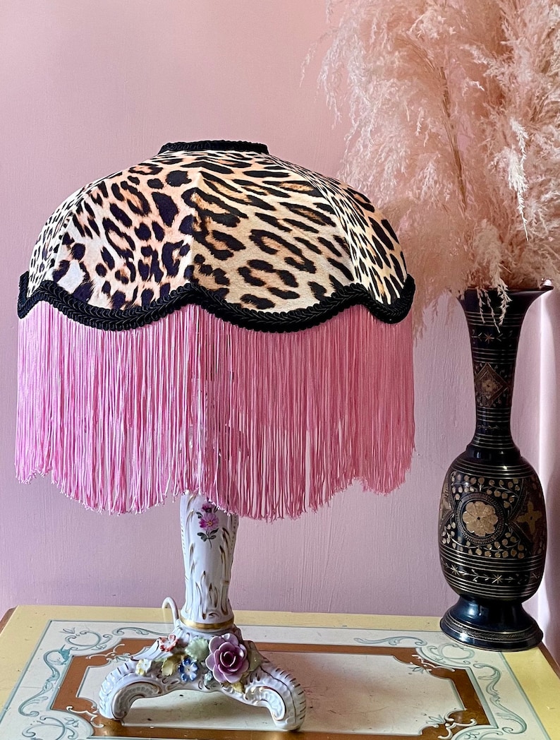 May include: A decorative table lamp with a leopard print lampshade, trimmed with black rope and pink fringe. The base is white porcelain with floral accents. A black vase with gold detailing is to the side.
