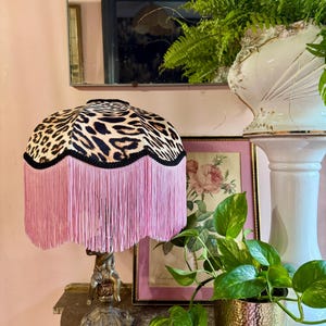 May include: A table lamp featuring a leopard print lampshade with pink fringe. The lamp has a detailed bronze base. The lamp is on a wooden surface, with a framed floral print and potted plants in the background.