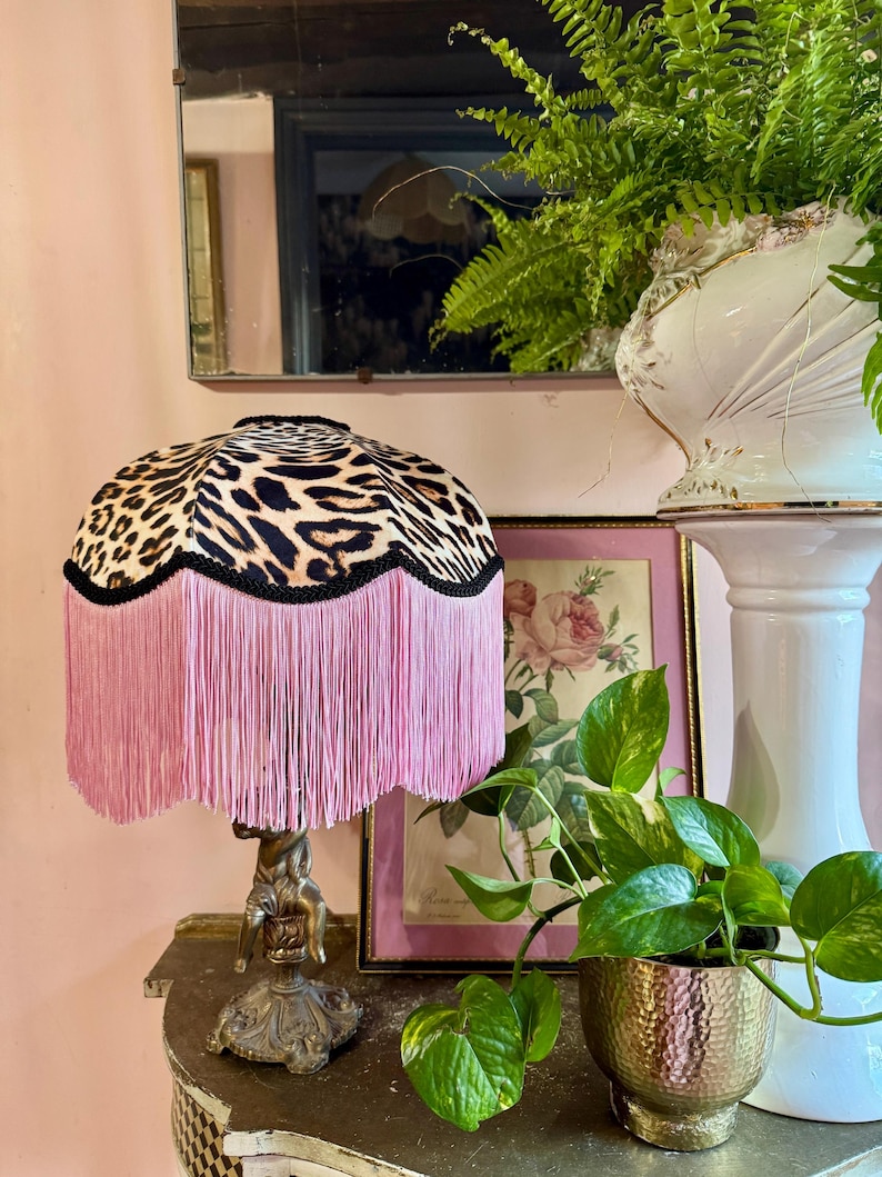 May include: A table lamp featuring a leopard print lampshade with pink fringe. The lamp has a detailed bronze base. The lamp is on a wooden surface, with a framed floral print and potted plants in the background.