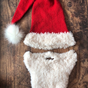 Könnte beinhalten: Eine rote gestrickte Weihnachtsmannmütze mit weißem Kunstfellbesatz und einem weißen Bommel oben. Ein weißer Kunstfellbart ist ebenfalls enthalten.