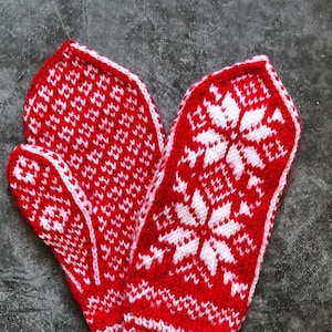 Könnte beinhalten: Ein Paar rote und weiße Strickhandschuhe mit einem Schneeflockenmuster. Die Handschuhe sind in der Mitte gefaltet und liegen auf einer grauen Oberfläche.