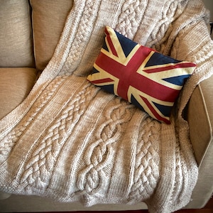 Peut inclure: Une couverture en tricot beige à motif torsadé est jetée sur un canapé. Un coussin rouge, blanc et bleu avec un motif Union Jack est posé sur la couverture.