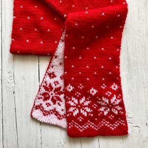 May include: Red knitted scarf with a white snowflake pattern. The scarf is folded in half and has a white border.