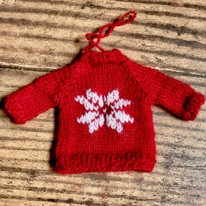 May include: A red knitted sweater ornament with a white snowflake design. The ornament has a loop for hanging.