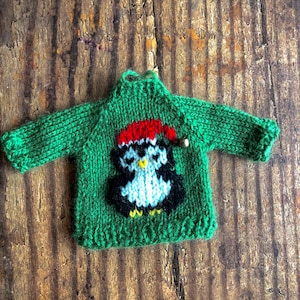 May include: A green knitted jumper with a penguin wearing a red hat. The jumper is made with a chunky yarn and has a simple design.