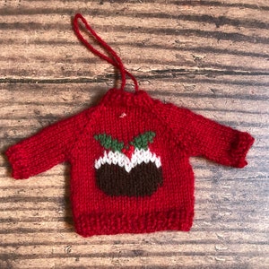 May include: A red knitted Christmas bauble shaped like a jumper with a brown and white Christmas pudding design on the front.