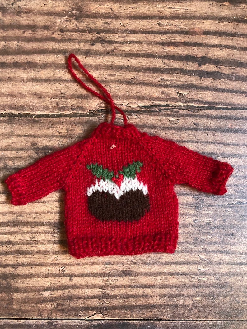 May include: A red knitted Christmas bauble shaped like a jumper with a brown and white Christmas pudding design on the front.