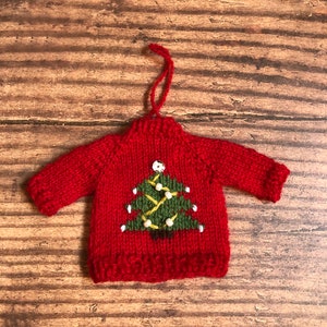 May include: Red knitted Christmas ornament shaped like a sweater with a green Christmas tree design on the front. The tree has white lights and a yellow star on top.