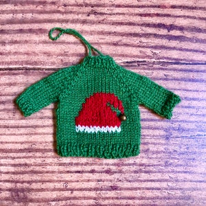 May include: Green knitted sweater ornament with a red and white Santa hat design. The ornament has a small gold bell.