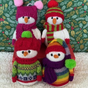 May include: Four knitted snowman ornaments. Each snowman has a unique hat, scarf, and sweater. The hats are red, pink, and striped. The scarves are red, green, and purple. The sweaters are green, blue, and rainbow colored. The snowmen have orange carrot noses and black eyes.