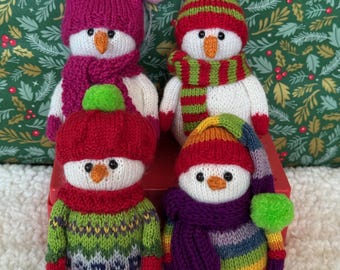 May include: Four knitted snowman ornaments. Each snowman has a unique hat, scarf, and sweater. The hats are red, pink, and striped. The scarves are red, green, and purple. The sweaters are green, blue, and rainbow colored. The snowmen have orange carrot noses and black eyes.