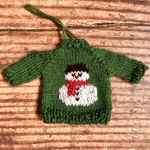 May include: Green knitted sweater ornament featuring a white snowman design. The snowman has a brown hat, red scarf, and white buttons.