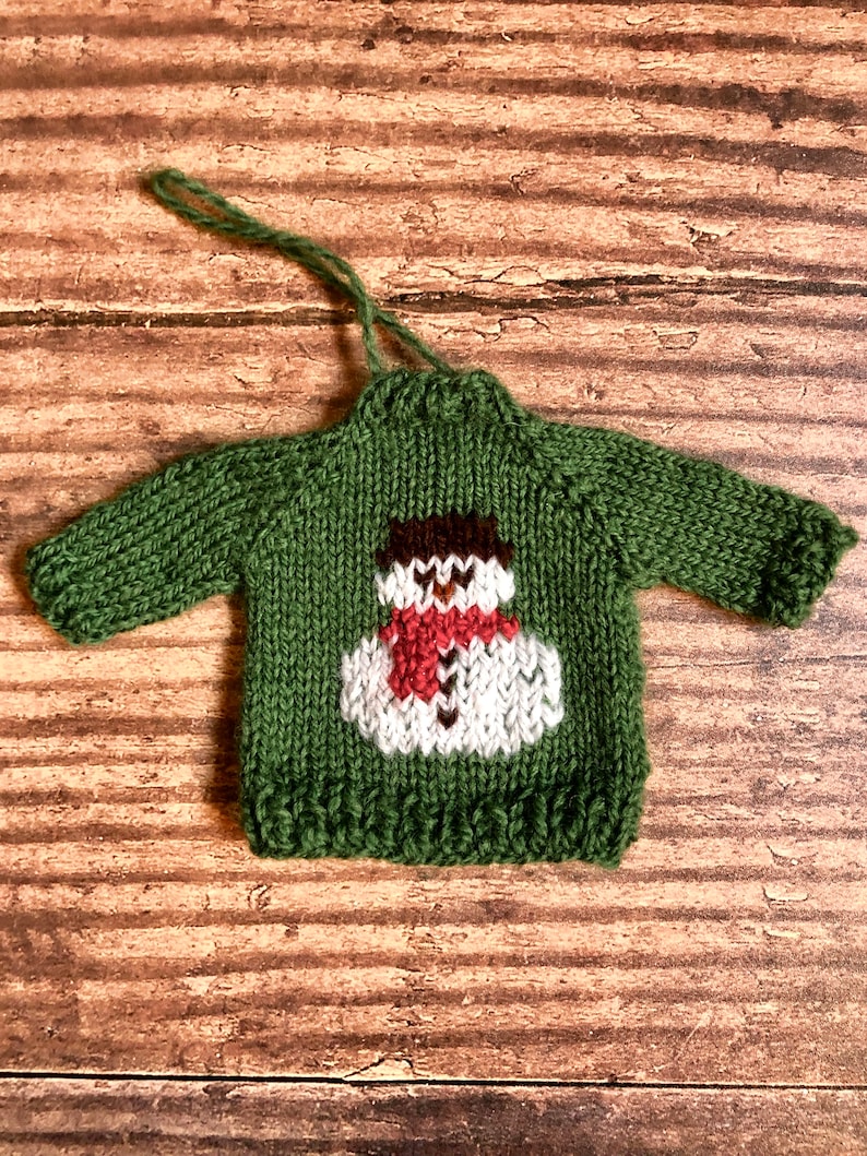 May include: Green knitted sweater ornament featuring a white snowman design. The snowman has a brown hat, red scarf, and white buttons.
