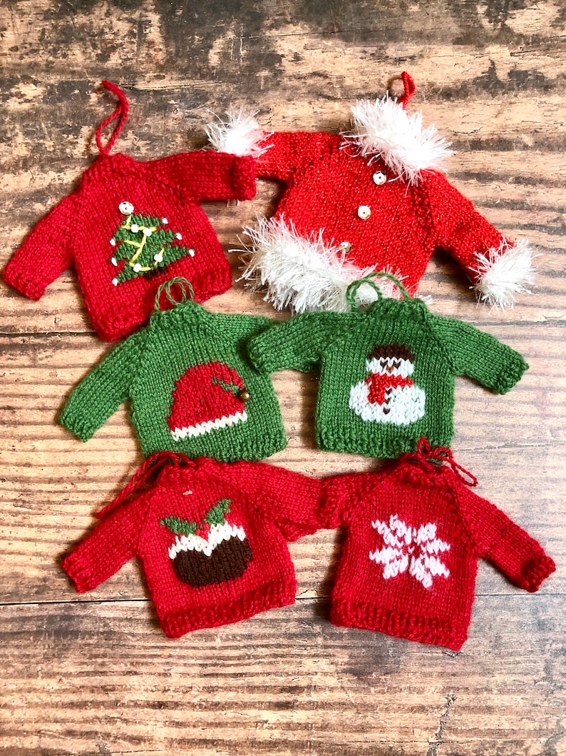 May include: Six knitted Christmas decorations shaped like jumpers with different festive designs. The jumpers are red and green with white accents. The designs include a Christmas tree, Santa Claus, a snowman, a Christmas pudding, and a snowflake.