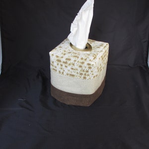 May include: A square tissue box cover with a brown, beige, and white patterned fabric. The top of the box has a round opening for tissues and is decorated with a brown fabric.
