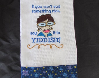 May include: White kitchen towel with blue and yellow stars at the bottom. The towel has an embroidered image of a woman with glasses and the text "If you can't say something nice, say it in YIDDISH!"