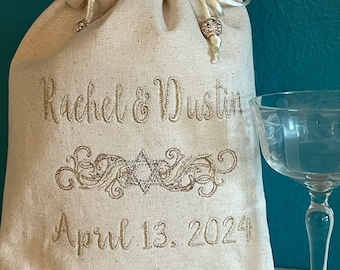 May include: A white fabric bag with a drawstring closure and gold embroidery. The embroidery includes the names "Rachel & Dustin" and the date "April 13, 2024". A gold star of David is centered between the names and date.