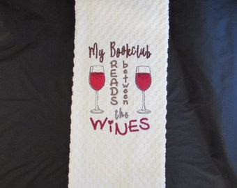 May include: White kitchen towel with brown embroidery that reads "My Bookclub Reads Between the Wines" with two red wine glasses.