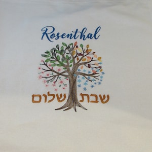 Puede incluir: Tela blanca con un árbol bordado en azul con hojas y flores de diferentes colores. El texto "Rosenthal" está bordado en azul sobre el árbol y el texto "עץ שלום" está bordado en oro debajo del árbol.
