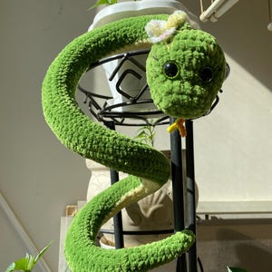 Pode incluir: Um brinquedo de cobra de crochê verde com uma textura macia e felpuda. A cobra apresenta olhos grandes, escuros e brilhantes, uma pequena língua amarela e um delicado acessório de flor branca e rosa preso no topo da cabeça.